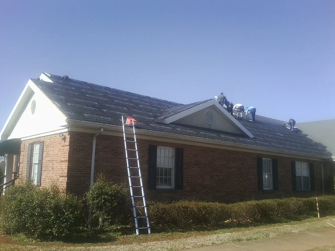 Crew tearing shingles off a brick ranch roof with a ladder set — Emergency Roof Repair work by our crew in Brightwaters