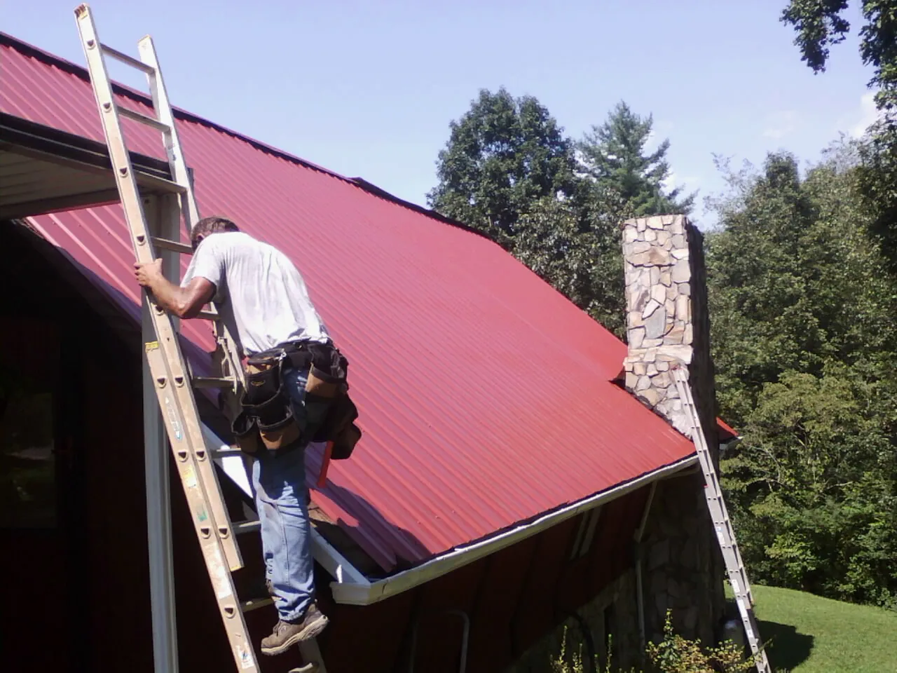 Roofer on a ladder beside a red metal roof and stone chimney — Soffit & Fascia Repair by Roofing Brothers in Brightwaters